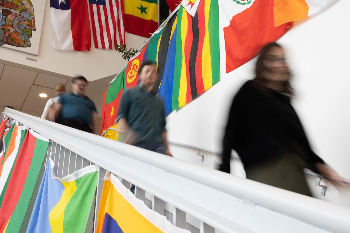 Three blurred students walk down a stairway past flags from many countries.
